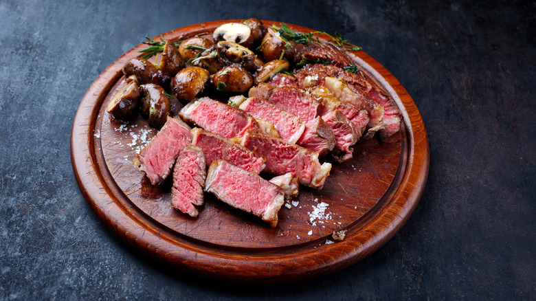 Dry-aged steak slices on circular wooden board, with mushrooms and herbs next to them