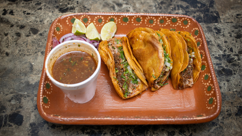Overhead view of birria tacos on a tray, with limes, onions, and cup of sauce
