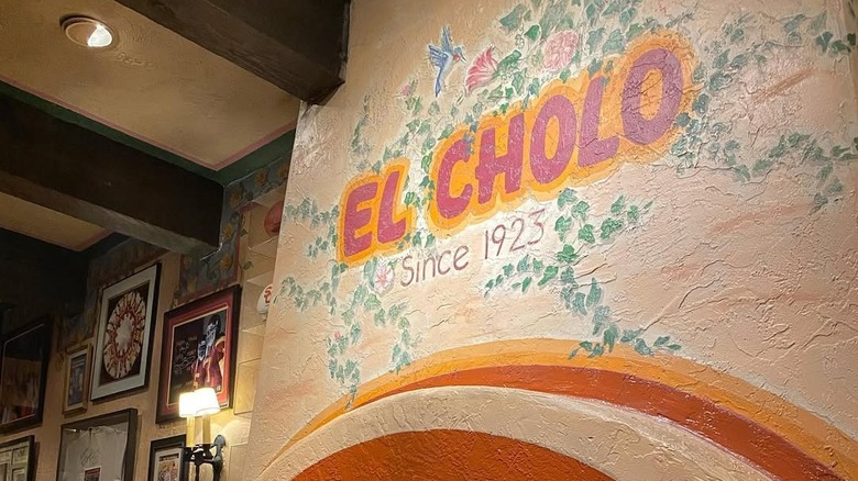 Restaurant interior with a colorful painted sign on the wall saying El Cholo, since 1923, and some framed pictures.