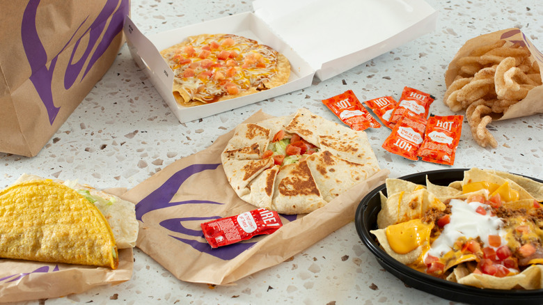 A feast of Taco Bell items, including a crunchy taco, a crunchwrap supreme, hot sauce, a Mexican pizza, Nachos Bell Grande, and Cinnamon Twists, next to a brown paper Taco Bell bag.