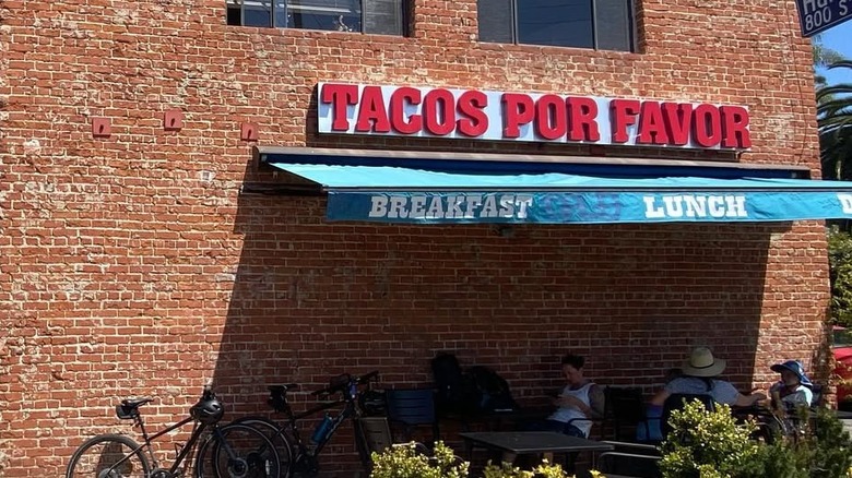 A brick building with a red sign reading Tacos Por Favor hanging over a green awning.