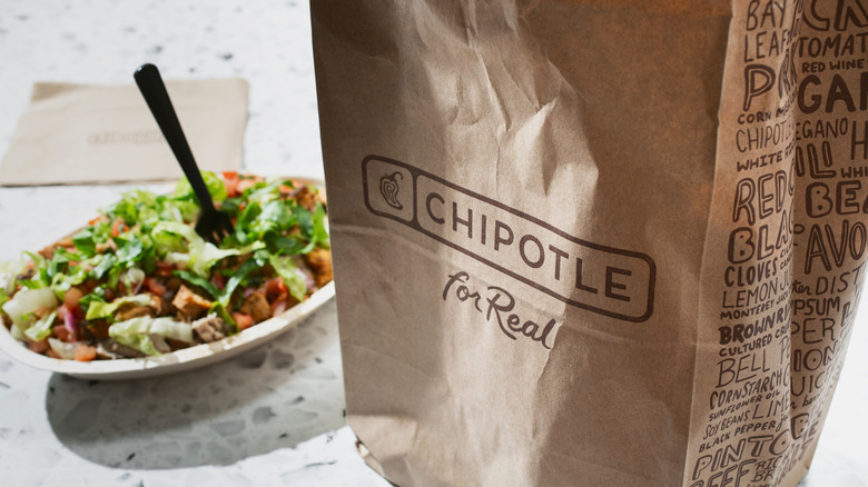 A burrito bowl next to a brown paper Chipotle bag.