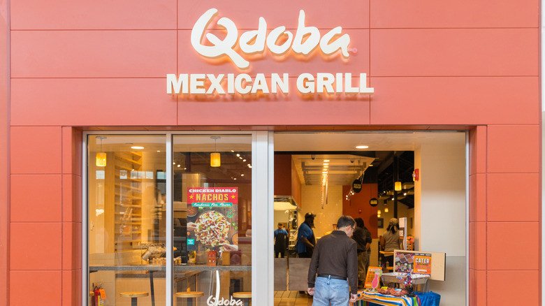 A customer walking in to a red-orange building with Qdoba Mexican Grill over the front door.