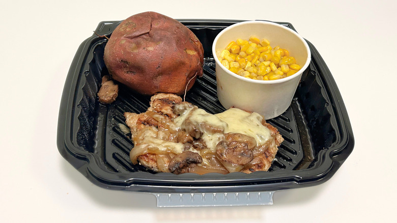 A black takeout tray on a white surface containing Smothered Chicken, sweet potato, and buttered corn