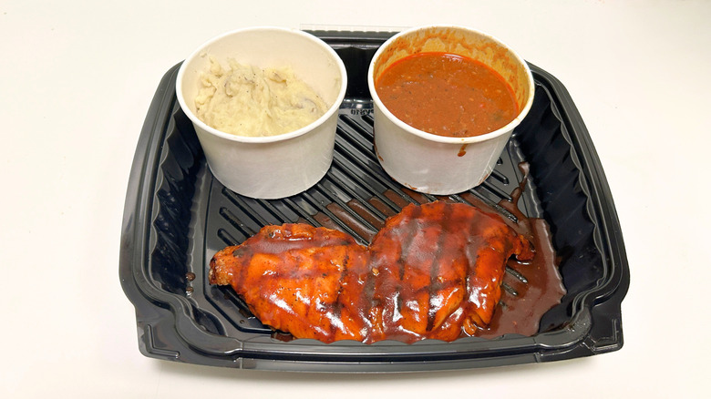 A black takeout tray on a white surface containing Grilled BBQ Chicken, mashed potatoes, and chili