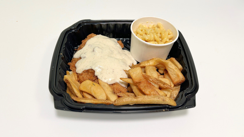 A black takeout tray on a white surface containing country fried chicken, steak fries, and mac and cheese