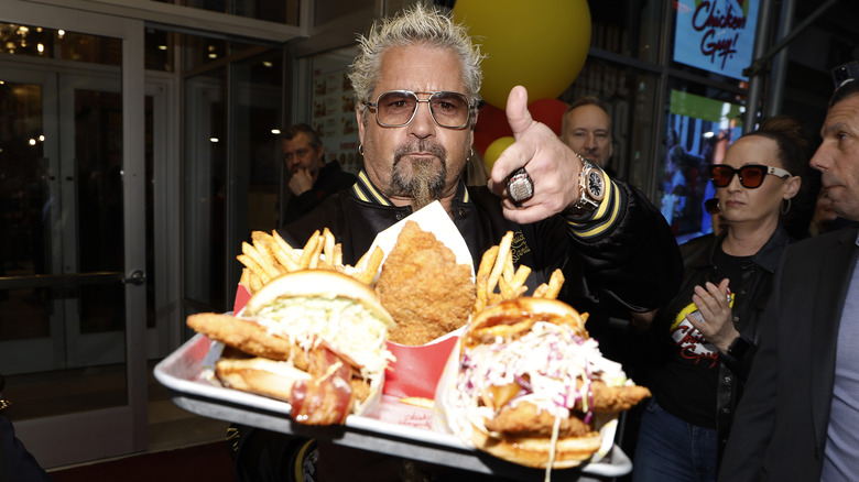Guy Fieri points to a tray of Chicken Guy foods including sandwiches, fries, and tenders