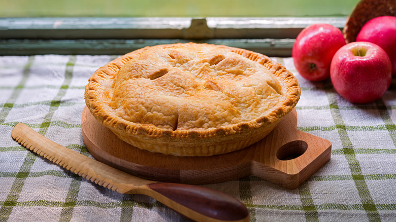 Pie on wood board next to wood knife and apples