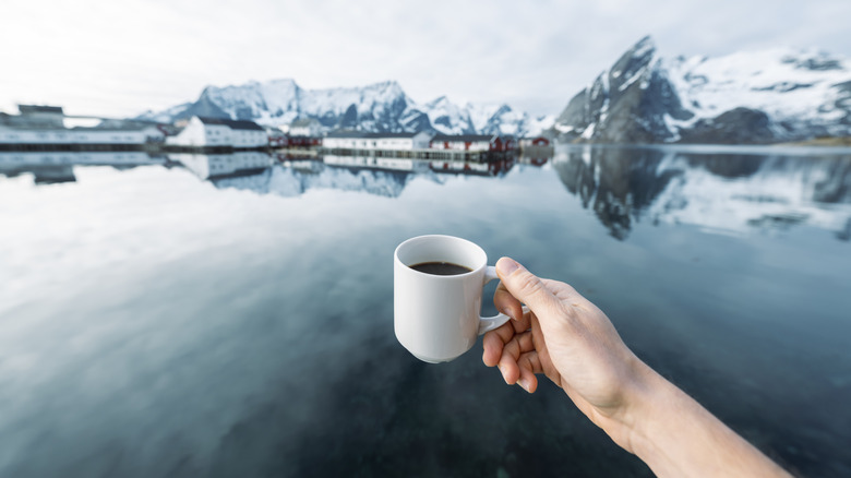 a hand holding up a cup of coffee with the background of a Nordic village and snowy mountains