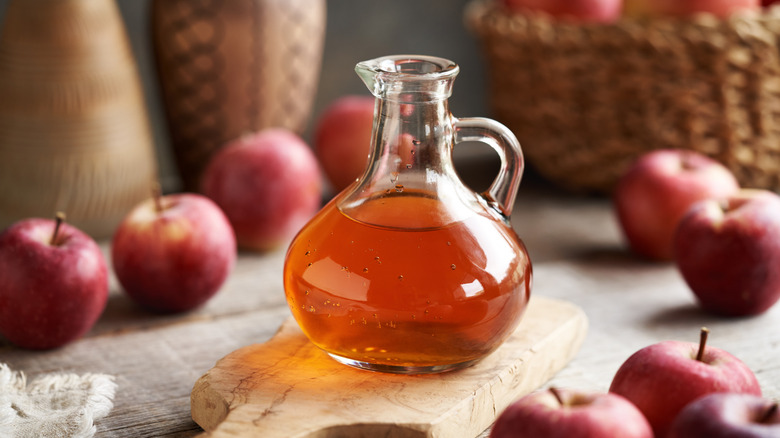 Jug of apple cider vinegar with apples strewn across a table with a basket in the background.