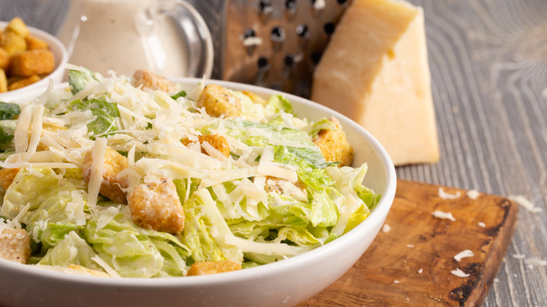 Caesar salad with dressing, croutons, and Parmesan in a white bowl and cheese and a grater in the background