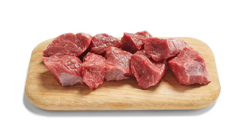 Cubed raw beef stew meat on a small wooden cutting board on a white background