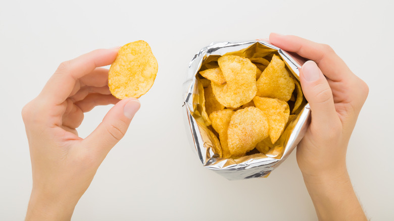 close-up overhead shot of a woman's hands holding a potato chip in one hand and open bag of chips in the other