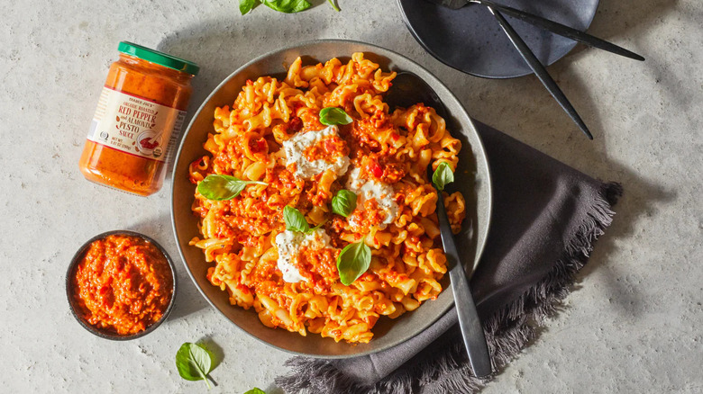 Large pasta bowl next to smaller bowl and jar of red pepper and almond pesto sauce