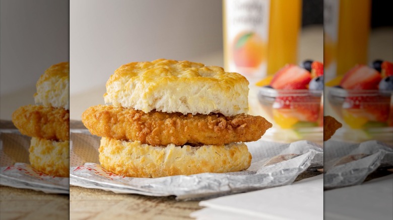 A Chick-fil-A chicken biscuit with a blurred cup of fruit and bottle of Simply orange juice in the background