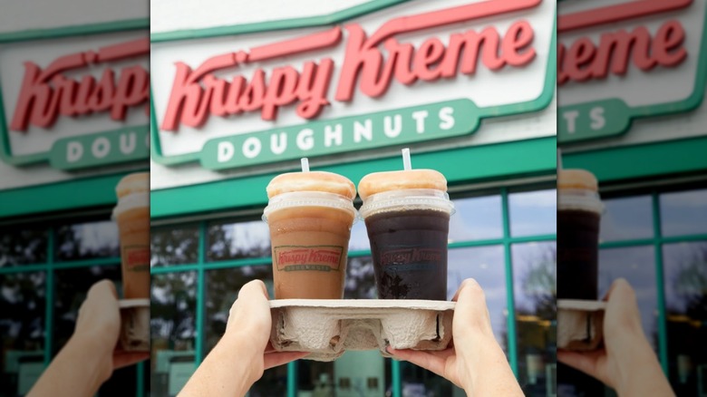 Krispy Kreme to-go drinks with glazed doughnuts on top being held up in front of a store sign
