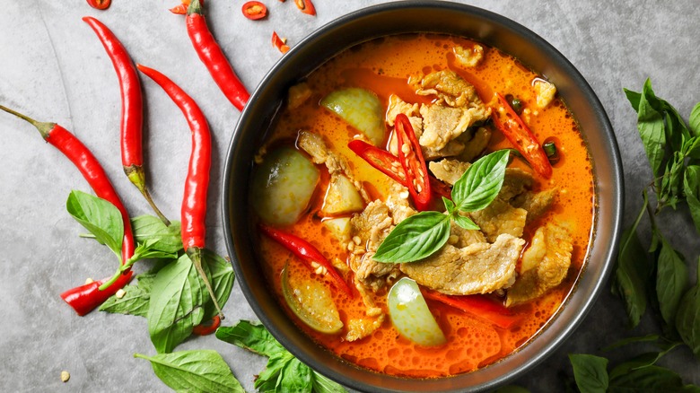 Thai red curry with pork in a bowl on table next to red chiles and basil leaves