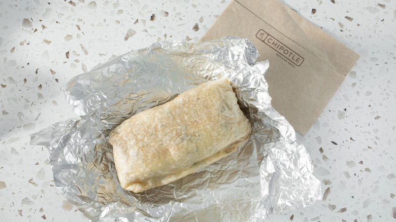 A burrito from Chipotle on a piece of aluminum foil, placed on a table with a Chipotle bag