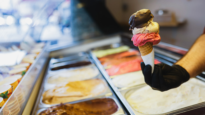 A hand holding a triple-stacked ice cream in a cone over an ice cream parlor freezer