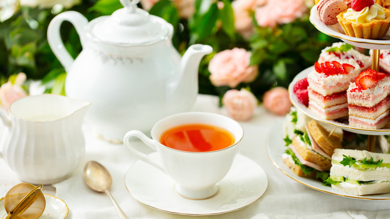 A tea pot, cup of tea, and a tiered stand of sandwiches and cakes