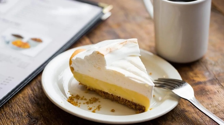 A slice of lemon meringue pie on a white plate with silver fork and white coffee mug