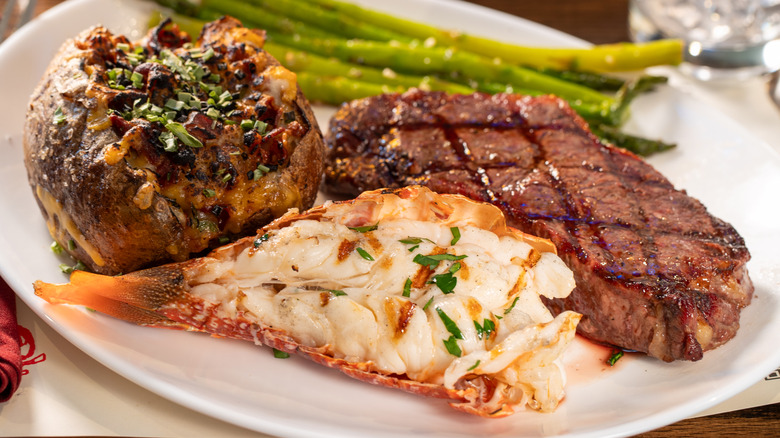 Plate of ribeye, lobster tail, stuffed baked potato, and asparagus at steakhouse.