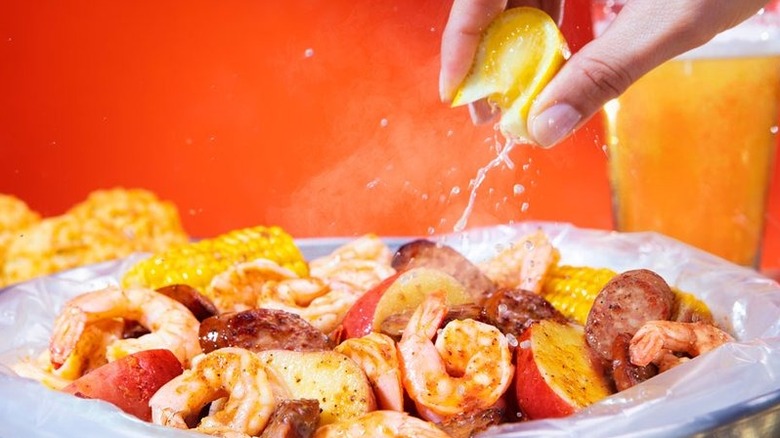 A person squeezing a lemon over a seafood boil of shrimp, sausage, corn  and potatoes