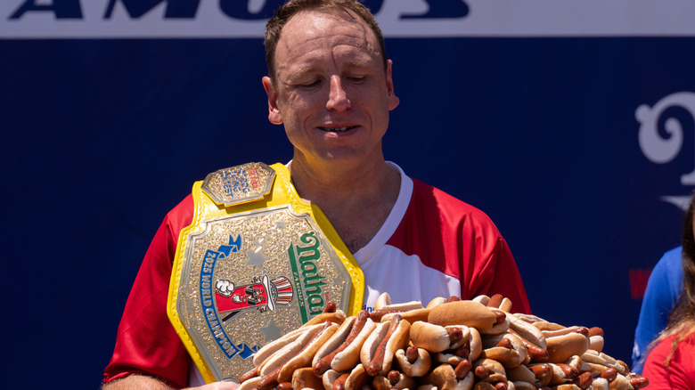 Joey Chestnut looks at pile of hot dogs, with belt on shoulder