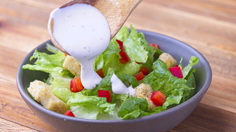 Wooden spoon pouring ranch dressing onto bowl of salad