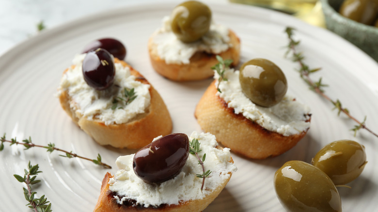 Olives and cream cheese tea sandwiches on a plate with sprigs of thyme and different colors of olives on top.
