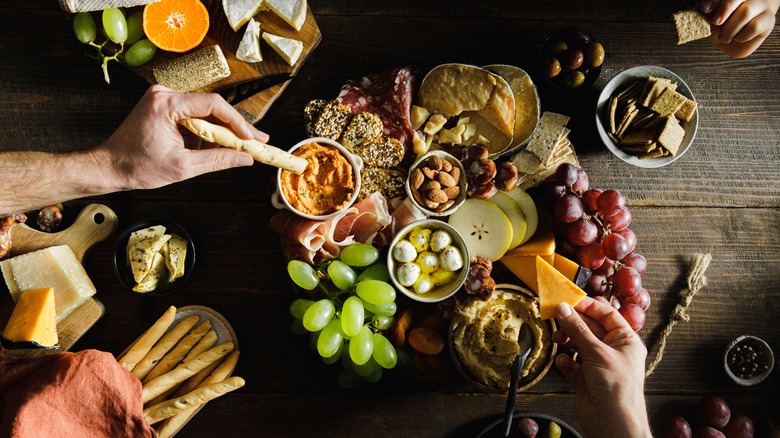 Hands digging into a charcuterie spread with cheese, meats, and fruits