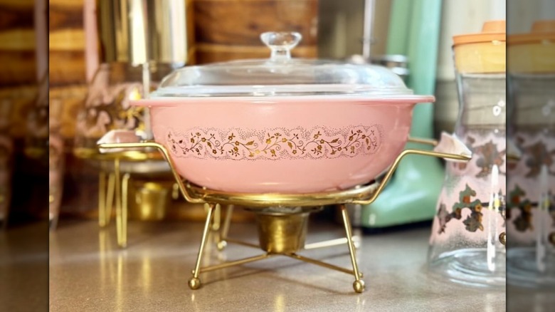 The Pink Pyrex Duchess casserole dish with clear lid sitting on a gold warming stand on a countertop