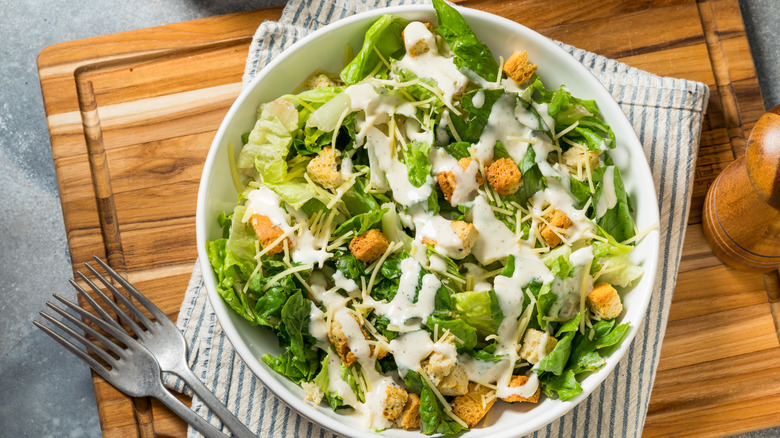Overhead view of Caesar salad in bowl with fork on a wood board.