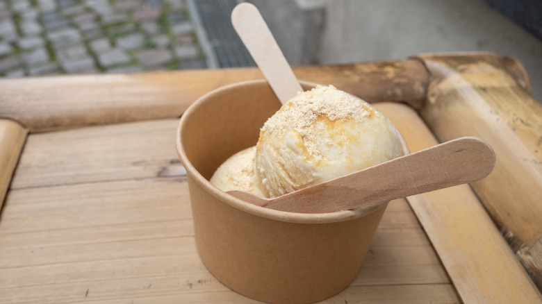 A small carboard cup of ice cream with two wooden spoons on it on a wooden surface outside