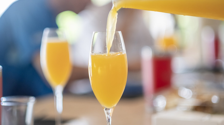 A bottle pouring mimosa into a champagne flute with a full flute in the background.