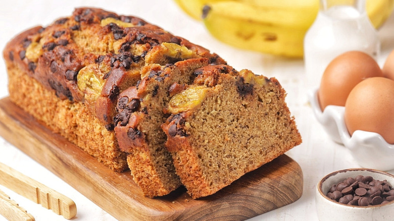 A loaf of banana bread on a cutting board with a few slices cut and ingredients next to it