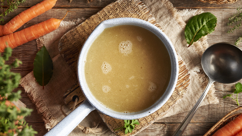 A small bowl of chicken stock, with a bay leaf and two carrots nearby