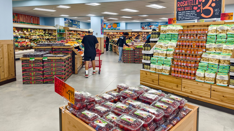 The interior of a Trader Joe's location in Florida with signs for plums and pasta sauce and a shopper with a red cart.