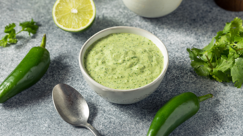 Peruvian green sauce in a white bowl surrounded by cilantro, limes, and jalapenos