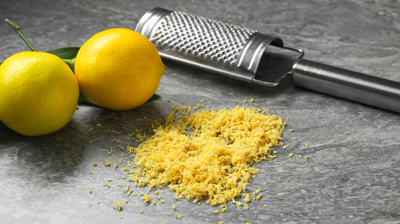 Lemon zest next to two lemons and a grater on a gray surface.