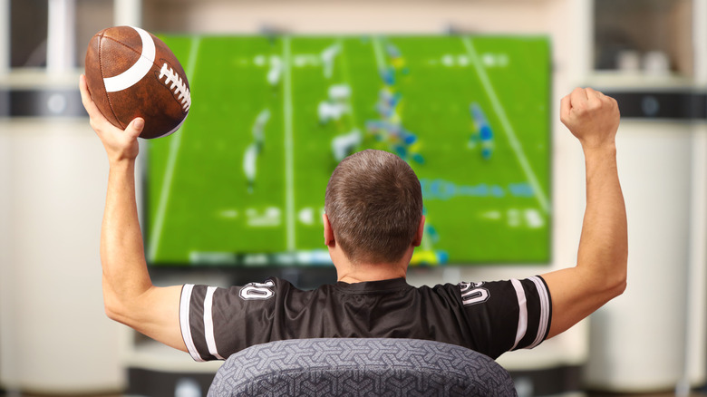 A man holding a football, while watching a game on TV, holding his arms up in celebration