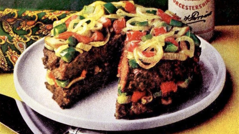 An old image of a burger layer cake topped with sauteed vegetables on a white plate