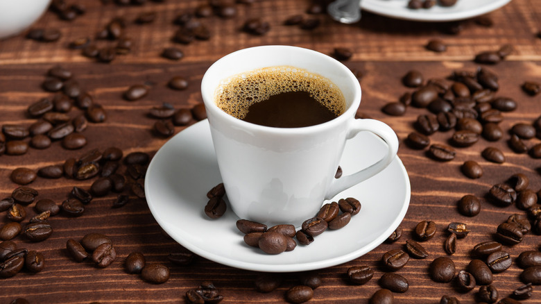 A small white mug of black coffee on a white plate with coffee beans scattered nearby
