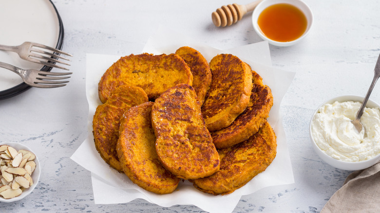 Pumpkin bread French toast slices on a plate with whipped cream, honey, and sliced almonds on the side