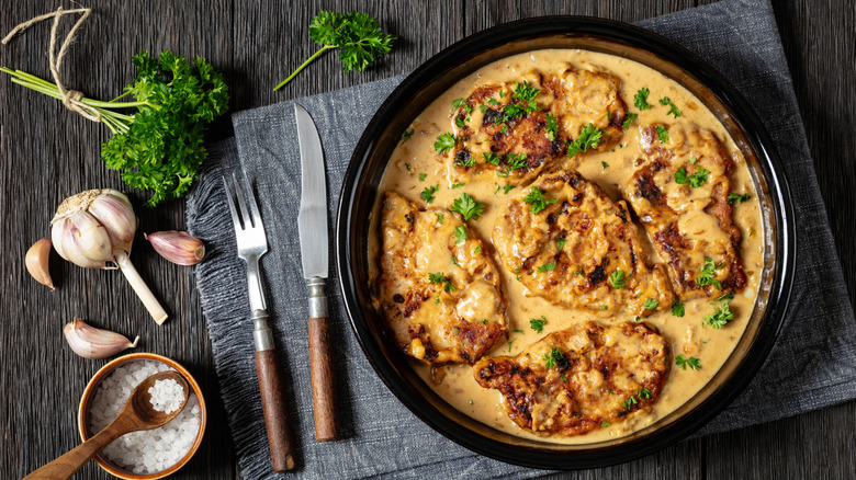 Smothered boneless pork chops are in a light brown gravy with chopped parsley.