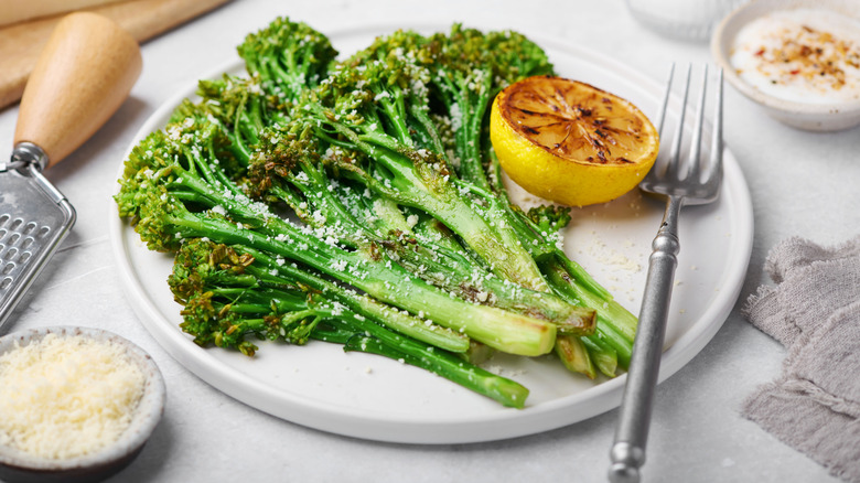 Broccolini that's been cooked and seasoned on a white plate with a lemon and fork on it.