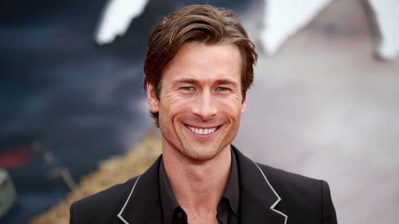 Glen Powell, shot from the shoulders up, smiling and wearing a black jacket