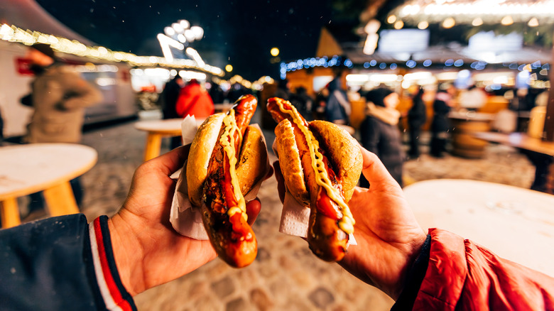 Two people holding hot dogs next to each other while out in a pubic market