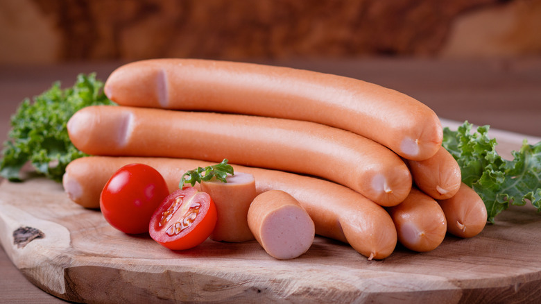 Six hot dogs stacked on a cutting board with tomatoes, lettuce and a sliced hot dog in front of them