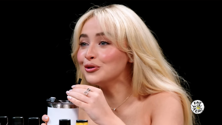 Sabrina Carpenter sits at the Hot Ones table, smiling and sipping a milkshake